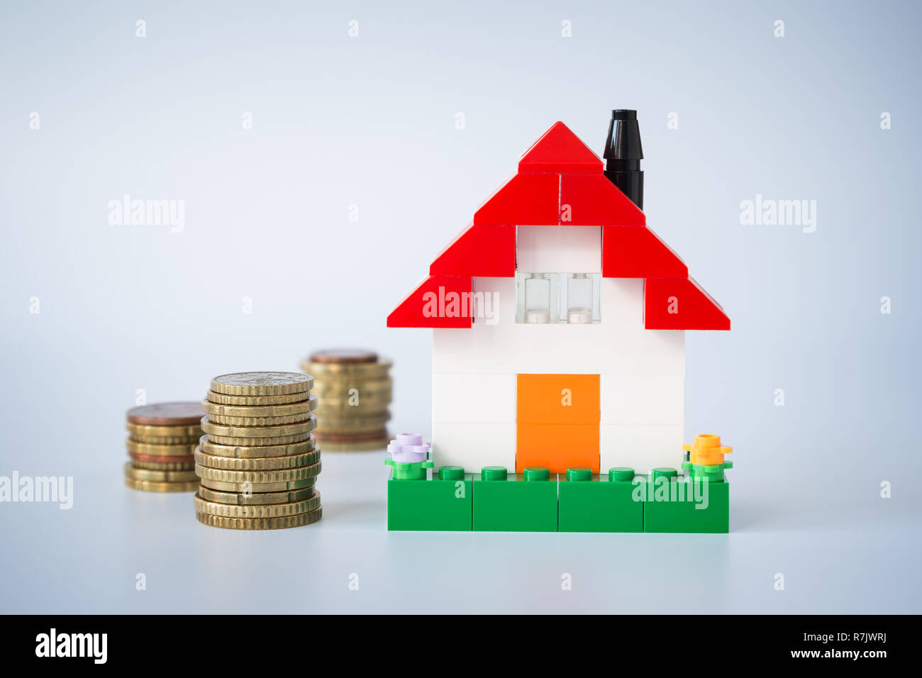 Stacks of coins and simple house made of toy building bricks, on gray background. Concept photo of mortgage, real estate business or house ownership. Stock Photo