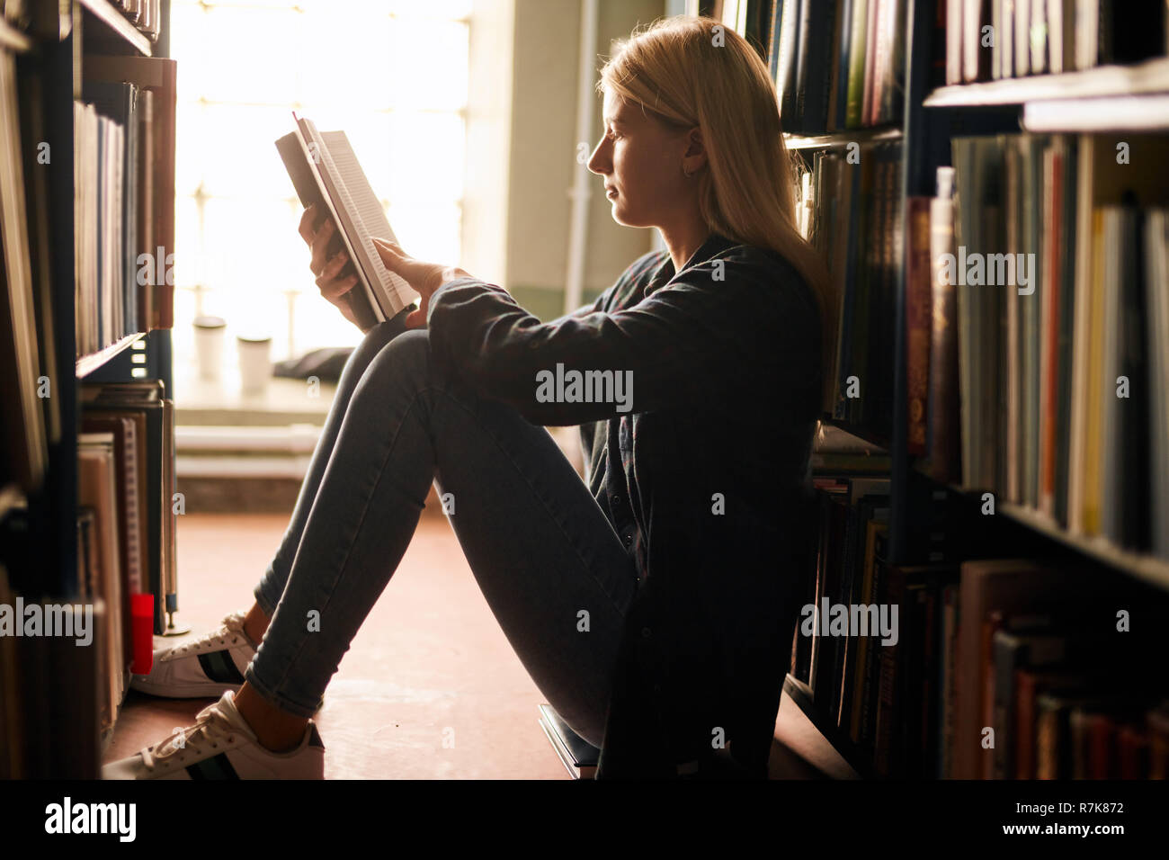 Young girl sitting on the floor in library between bookshelves, reading ...