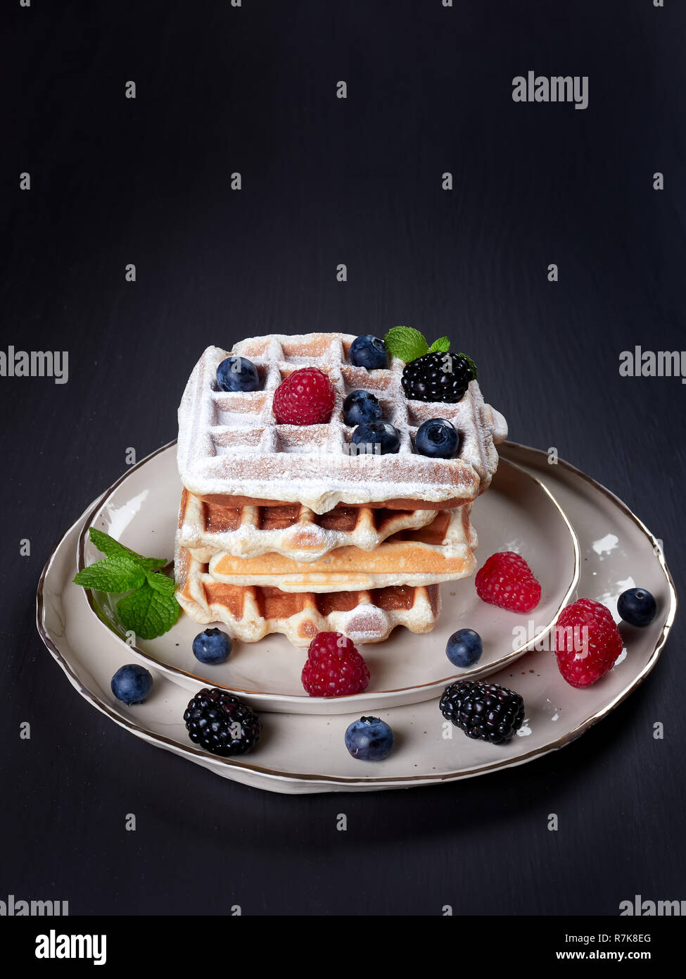 A stack of four home made Belgian waffles dusted with sugar, with fresh berries and mint leaves, on a beige platter, set on dark brown background Stock Photo