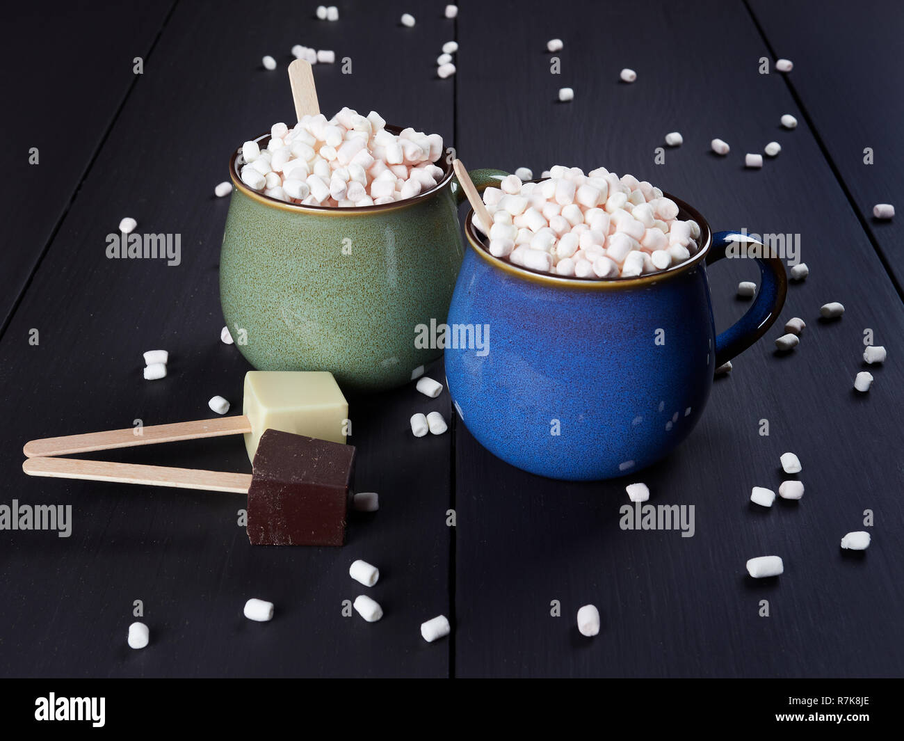Two cups with hot chocolate and mini marshmallows, one cup being blue and the other one green, with white and dark chocolate cubes, on dark brown back Stock Photo