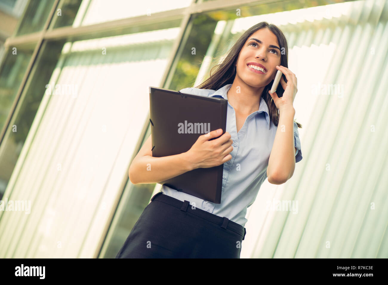 Young beautiful business woman with planner in her hands, standing in front of office building and talking on mobile phone Stock Photo
