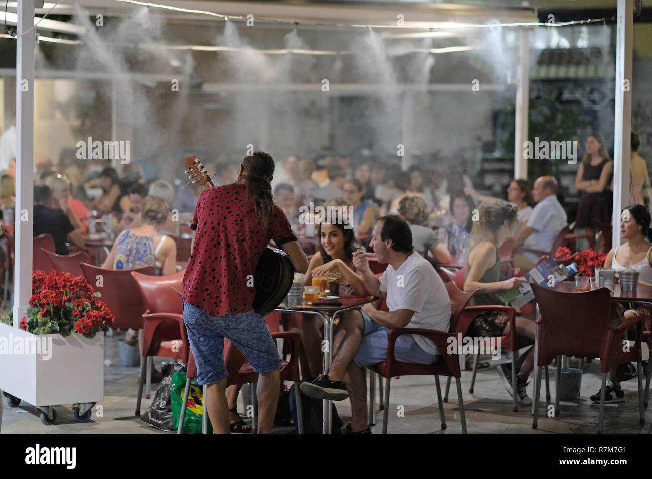 A musician-busker- plays to people in a Spanish bar with misters keeping the customers cool. Stock Photo