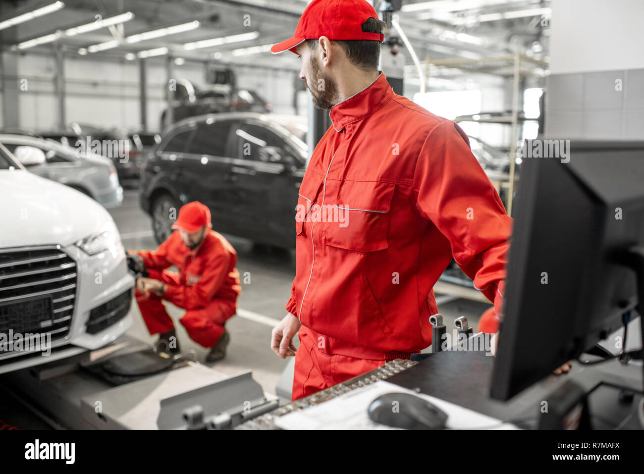 Two handsome auto mechanics in red uniform making wheel alignment with professional tools and computer at the car service Stock Photo