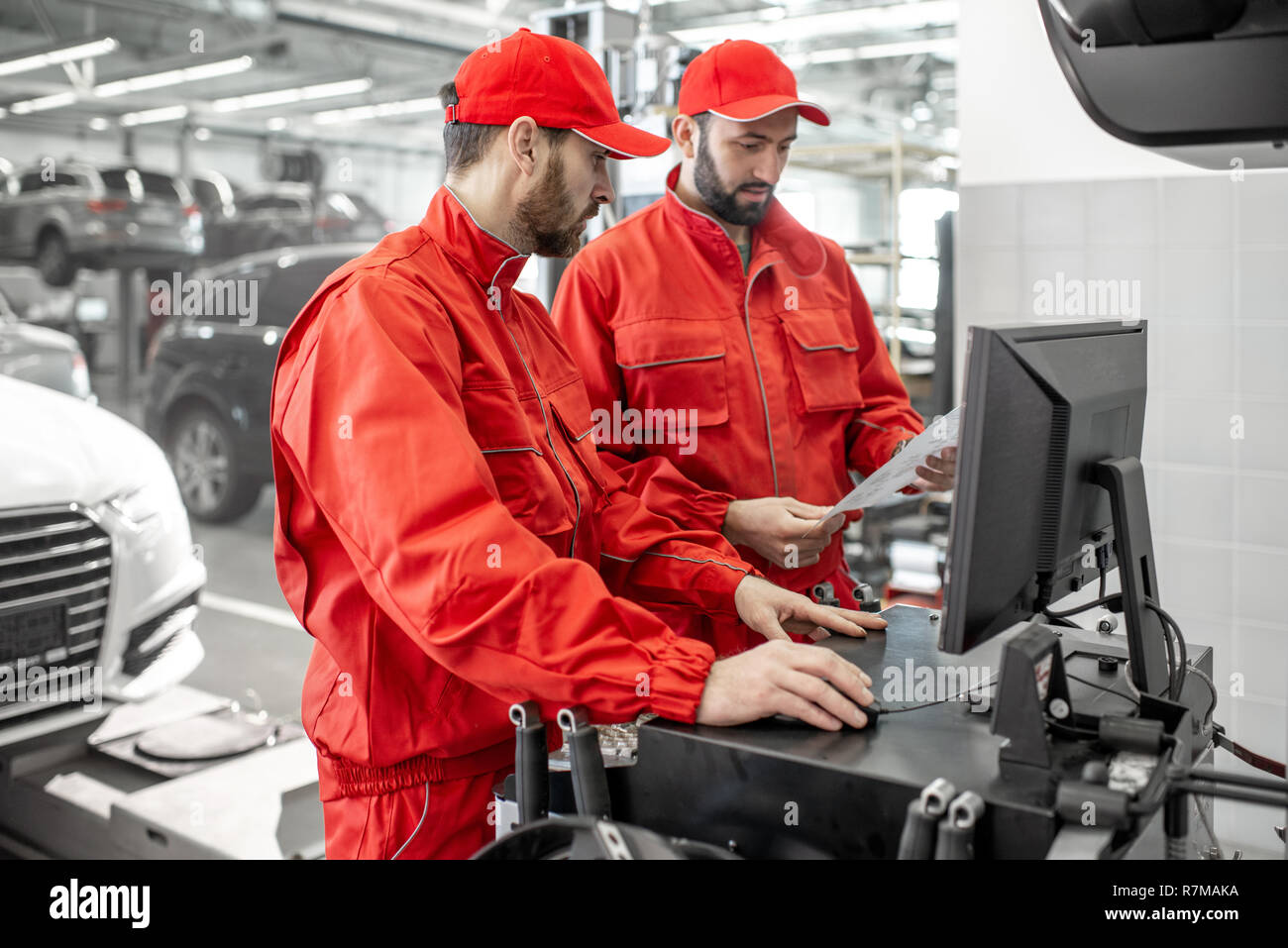 Two handsome auto mechanics in red uniform making wheel alignment with professional tools and computer at the car service Stock Photo