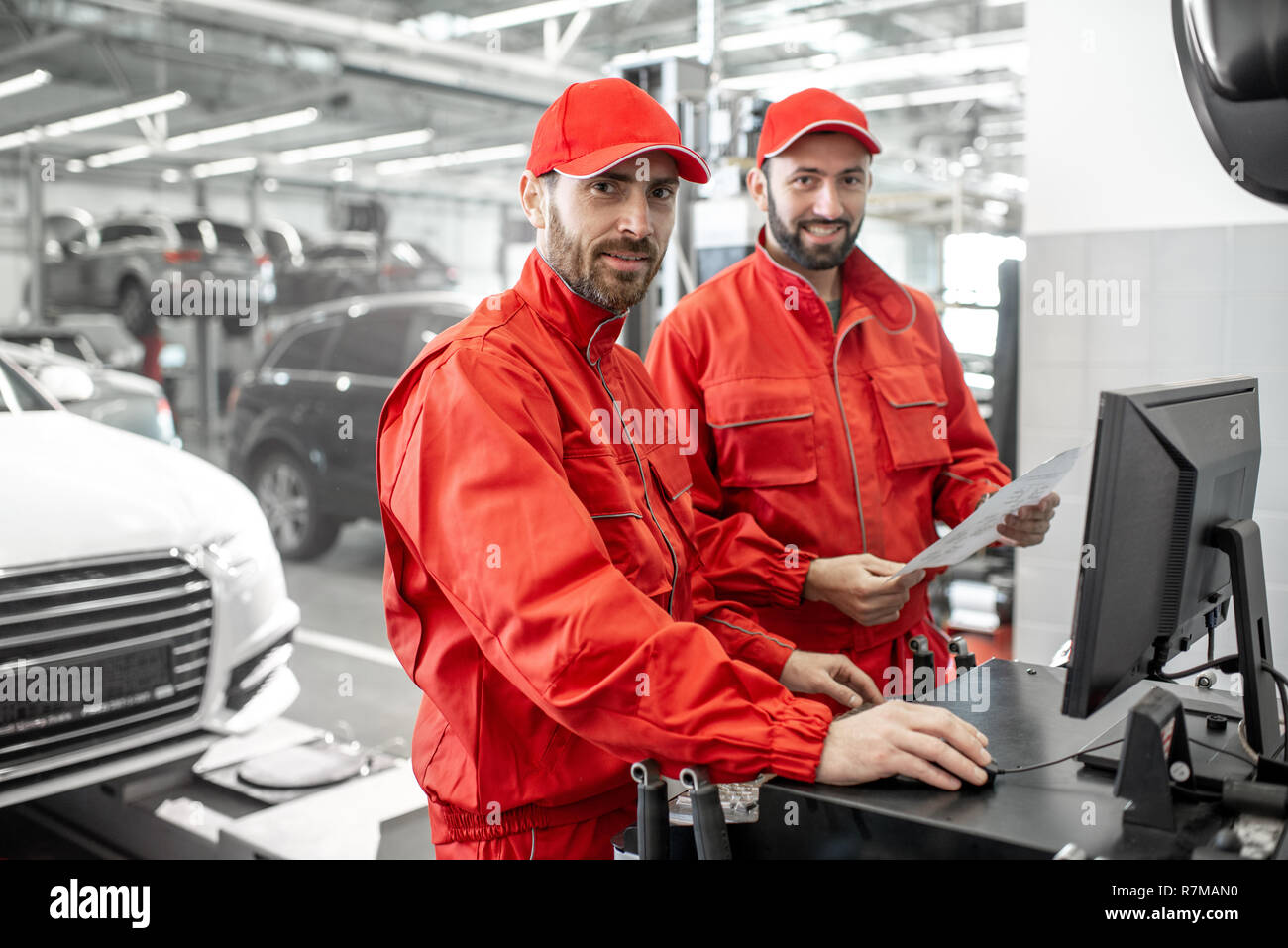 Two handsome auto mechanics in red uniform making wheel alignment with professional tools and computer at the car service Stock Photo
