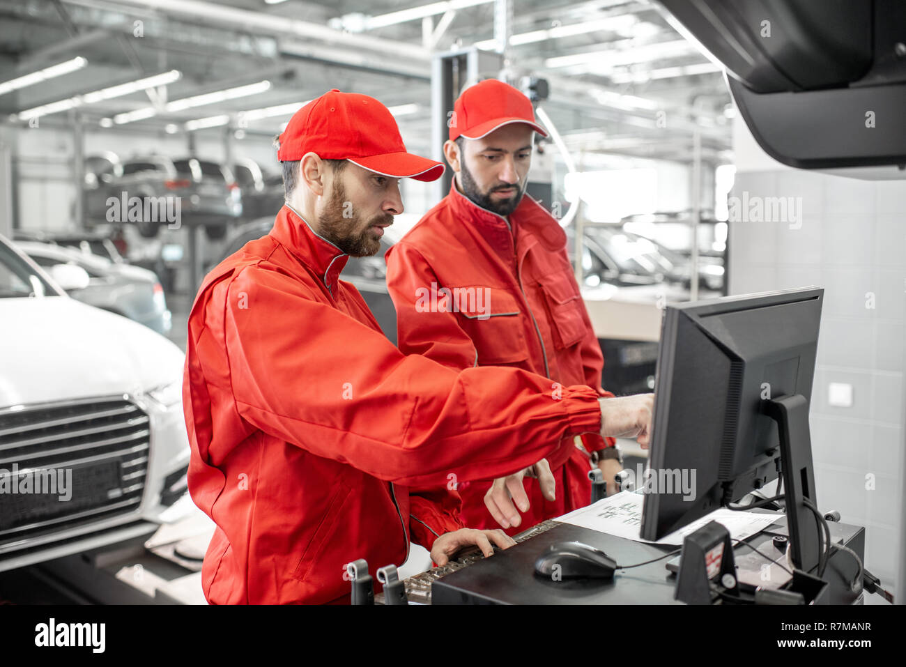 Two handsome auto mechanics in red uniform making wheel alignment with professional tools and computer at the car service Stock Photo