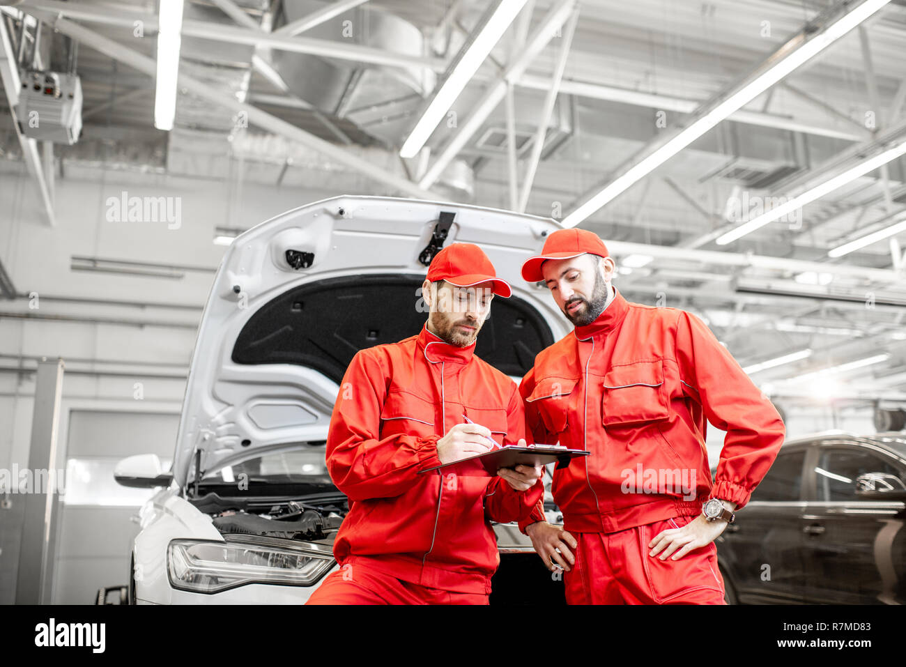 Two male auto mechanics in red uniform signing vehicle service documents standing near the car at the car service Stock Photo