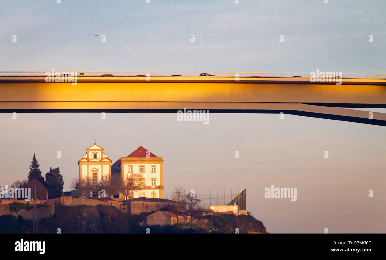 A church is seen at a distance, below the Ponte do Infante Bridge, in Porto, Portugal, during the golden hour, while seagulls fly by. Stock Photo