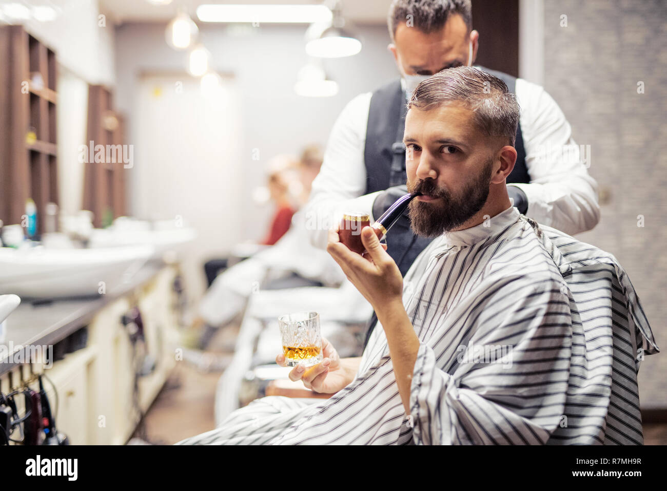 Hipster man client visiting haidresser and hairstylist in barber shop, smoking a pipe. Stock Photo