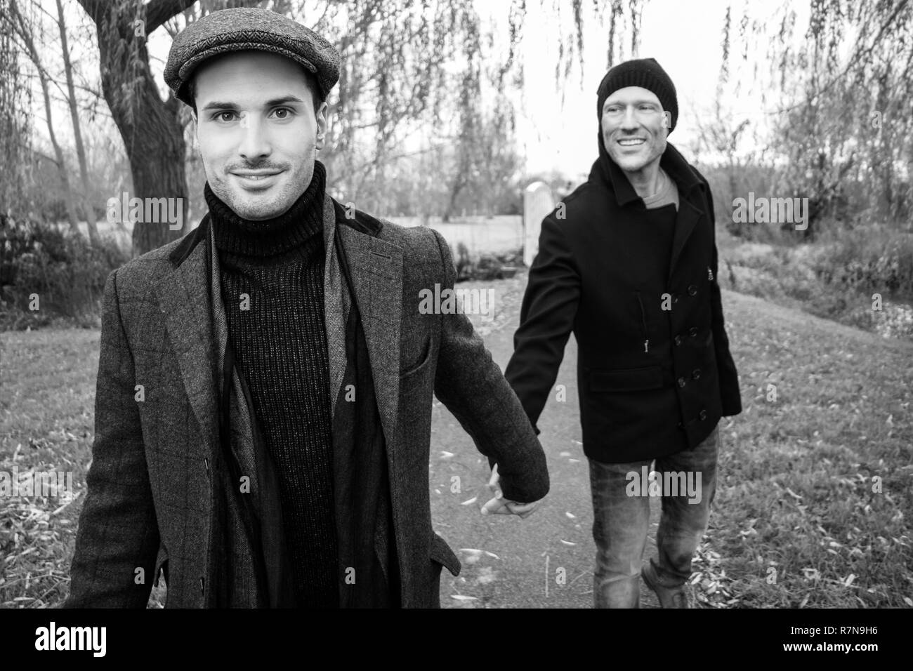 Black and white portrait of male gay couple holding hands and smiling as they walk on park pathway Stock Photo