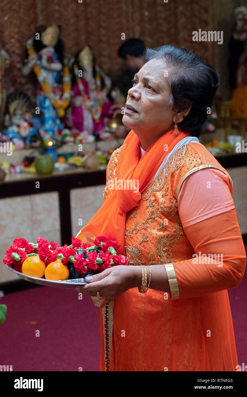 A devout Hindu woman prays in front of statues of the deities while holding a food offering. In New York City Stock Photo