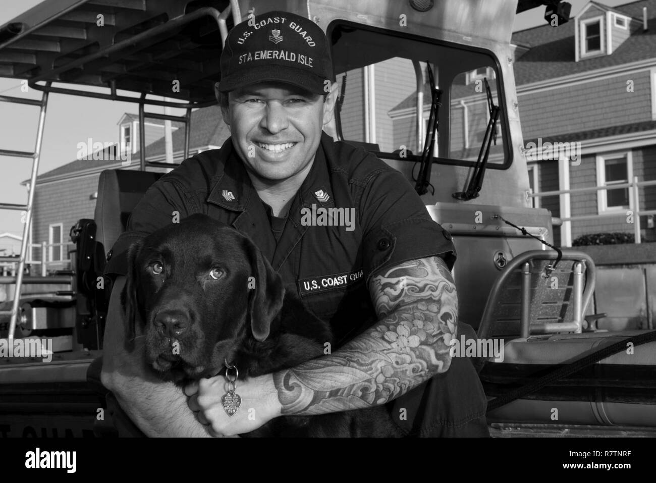 Coast Guard Petty Officer 1st Class Tyler Weeks, a machinery technician at Station Emerald Isle, North Carolina, poses for a portrait with his dog Boone near a 24-foot Special Purpose Craft-Shallow Water boat at the station, March 31, 2017. Boone is the station’s unofficial mascot and occasionally gets underway with the station’s boat crews. Stock Photo