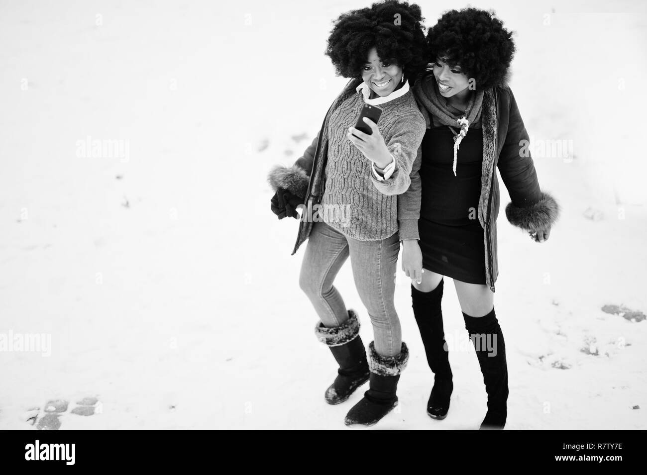 Two curly hair african american woman wear on sheepskin coat and gloves posed at winter day and making selfie on phone. Stock Photo