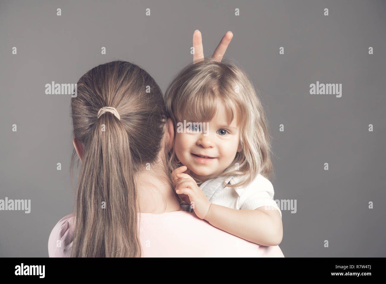 Young and slim mother stands with her back and holds in her arms a small wonderful child. Blonde adorable blue eyed baby girl is looking at the camera Stock Photo