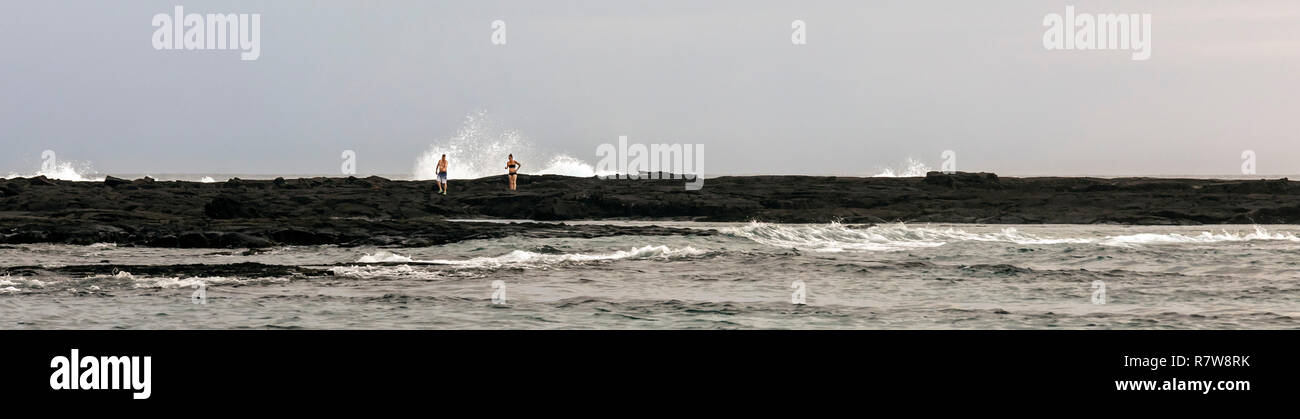 Honaunau, Hawaii - Waves break behind a man and a woman walking on rocks that extend into the Pacific Ocean on Hawaii's Big Island. Stock Photo