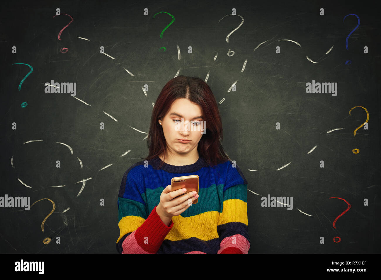 Emotional shock as cute young pretty woman standing over chalkboard background using mobile phone looking anxious and surprised try to solve different Stock Photo