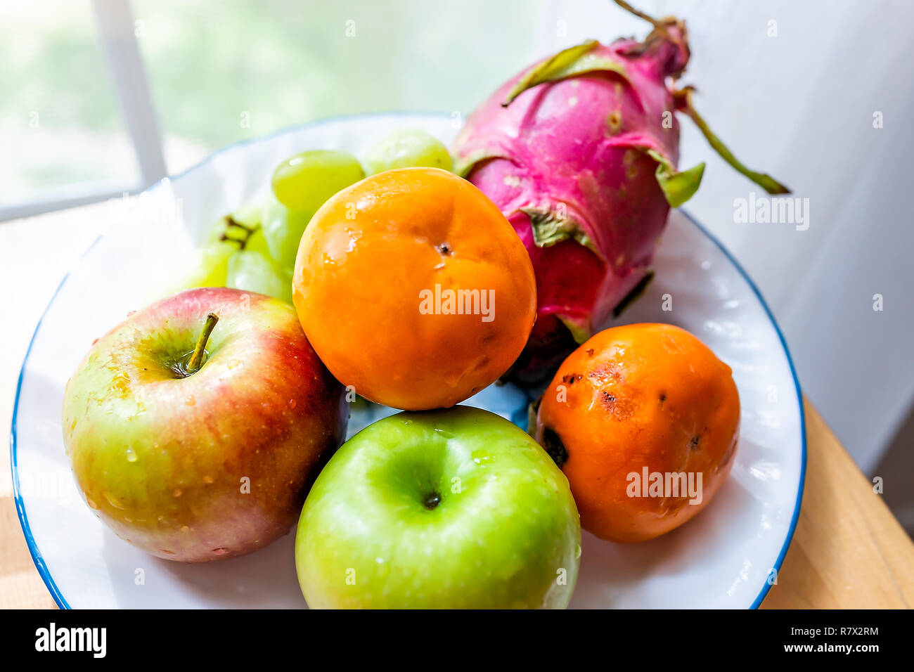 Closeup of whole orange, green, red dragon dragonfruit fruit fruits, apples, persimmons kaki, on plate, wooden table by window, autumn season Stock Photo
