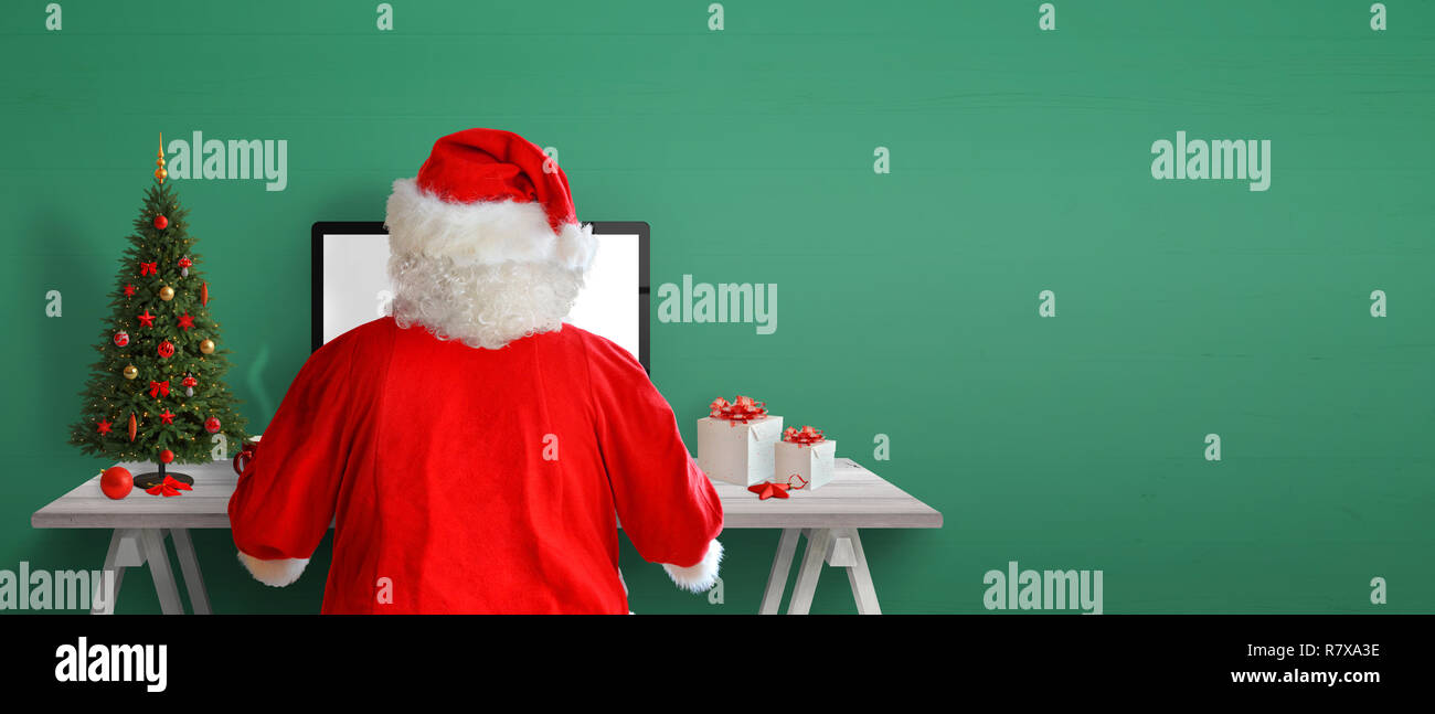 Santa Claus send letters online. Christmas tree, gifts and decorations on work desk. Green wall in background with copy space. Stock Photo