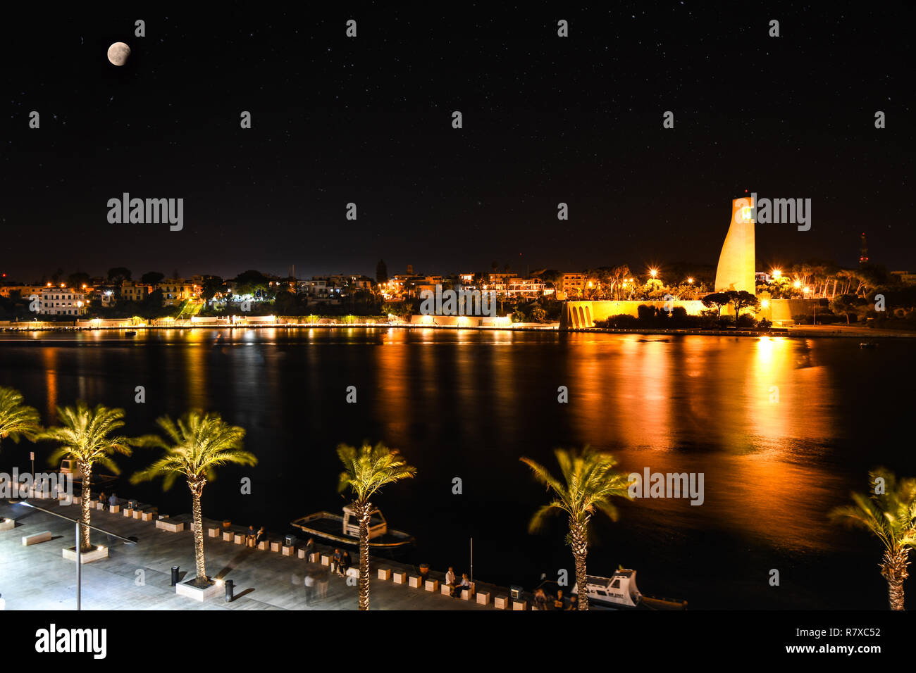 A full moon over the coastal port of Brindisi, Italy, as the Italian Sailors monument, bay and promenade are illuminated on a summer evening. Stock Photo