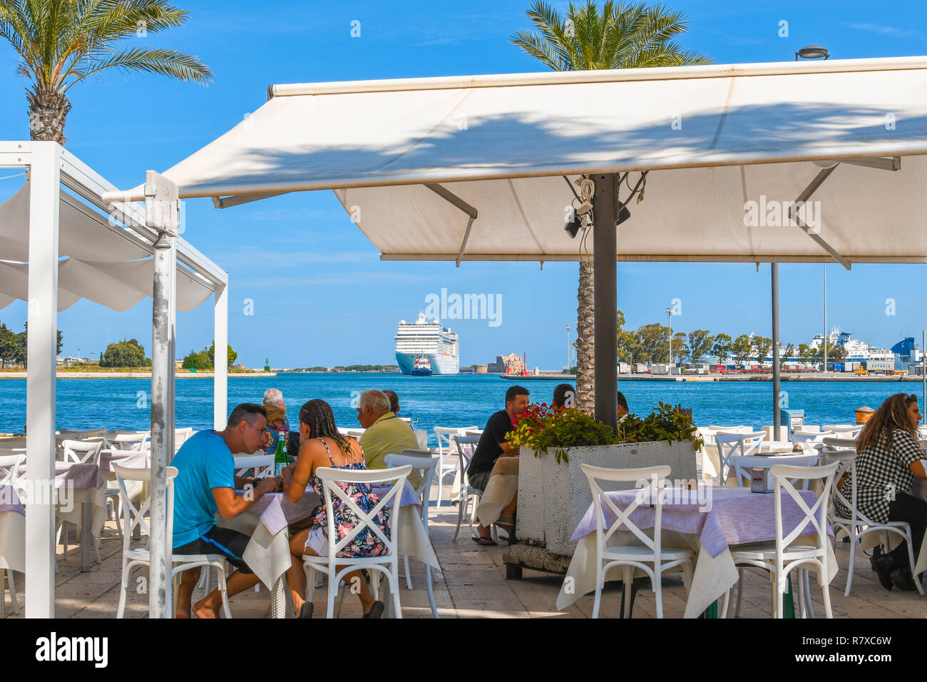 Tourists enjoy lunch at a waterfront cafe in the port city of Brindisi Italy as a massive cruise ship is towed into port. Stock Photo