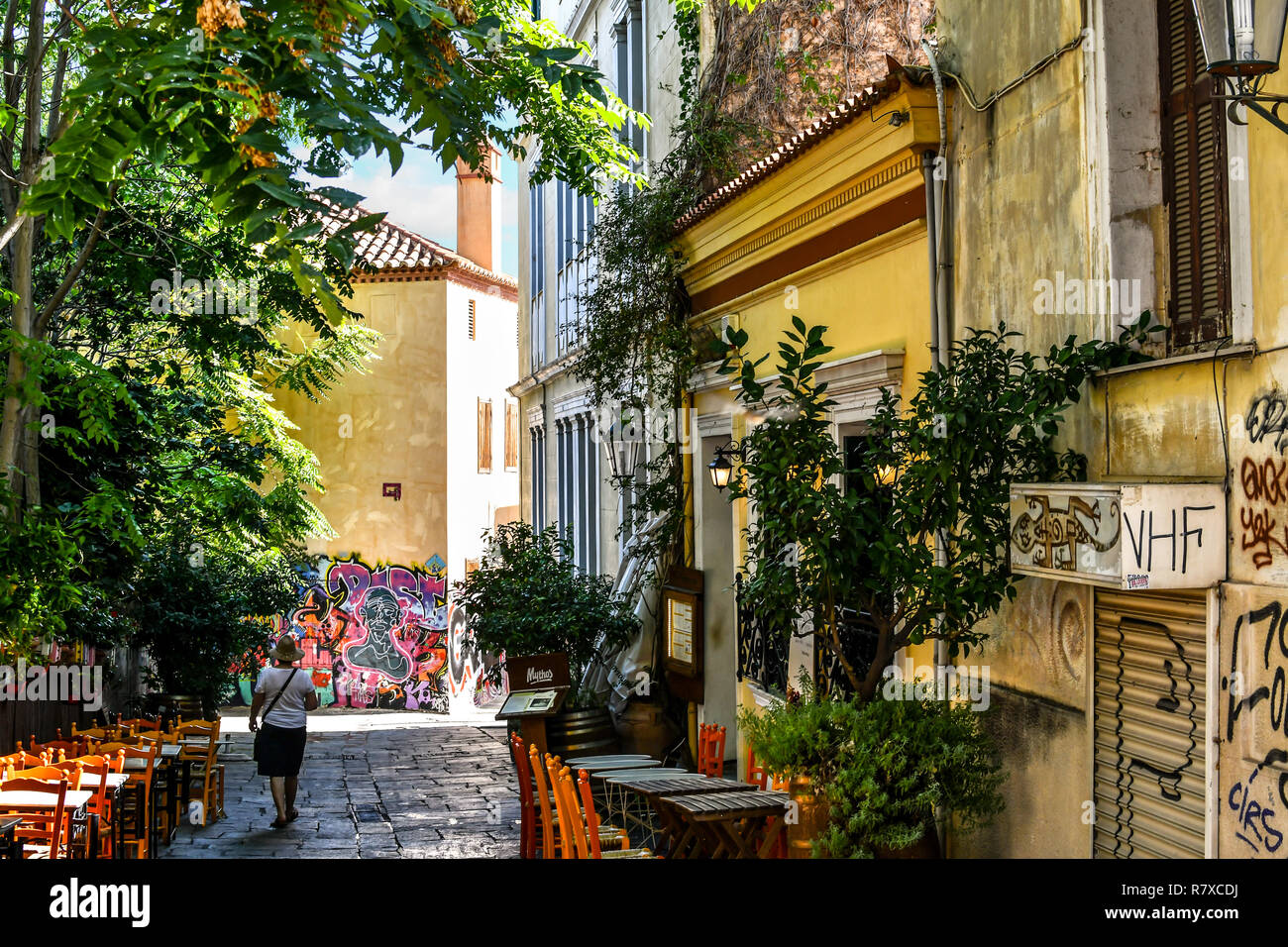 A solo female tourist walks past a small cafe with outdoor tables on a narrow shaded alley in the Plaka district of Athens, Greece Stock Photo