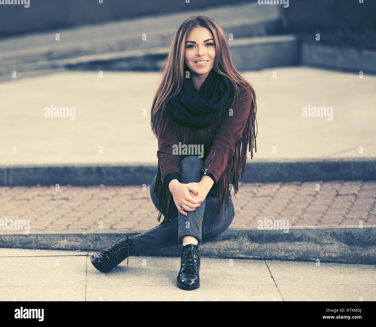 Happy young woman sitting on sidewalk in city street. Stylish fashion model in leather fringe suede jacket and blue jeans Stock Photo