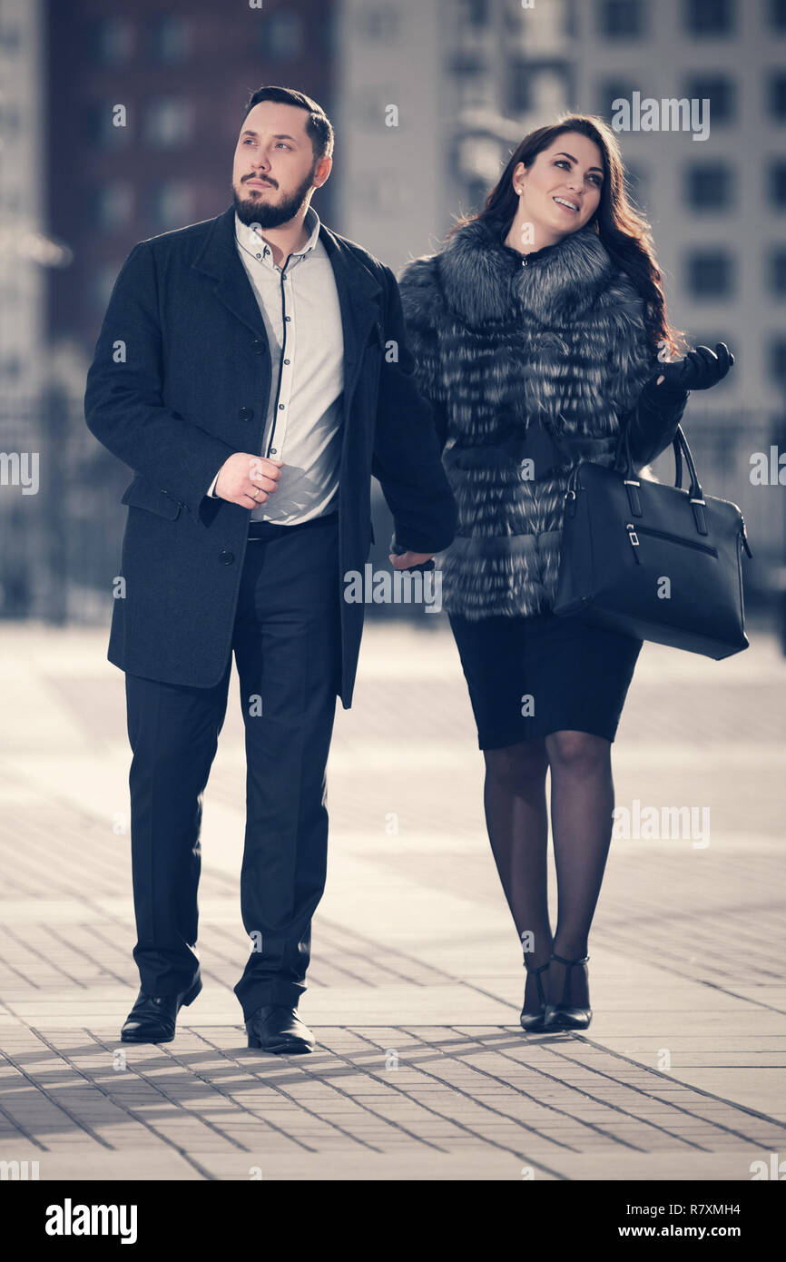 Happy young fashion couple walking on city street Stylish trendy man and woman wearing fox fur jacket and classic black coat Stock Photo
