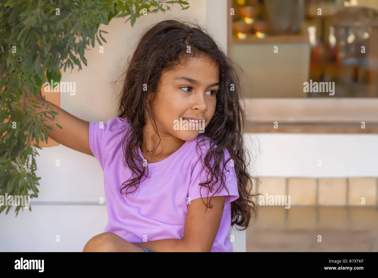 Beautiful little girl holds on to a branch while leaning on a wall looking away. Wearing a solid color light purple shirt with shabby chic messy hair. Stock Photo