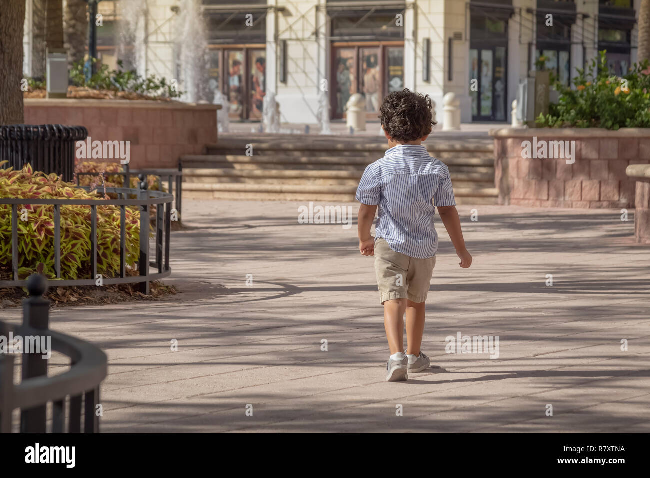 A confident little boy walks away across the courtyard on a sunny summer day. Dressed in tennis shoes, khaki shorts, and a blue striped shirt. Stock Photo