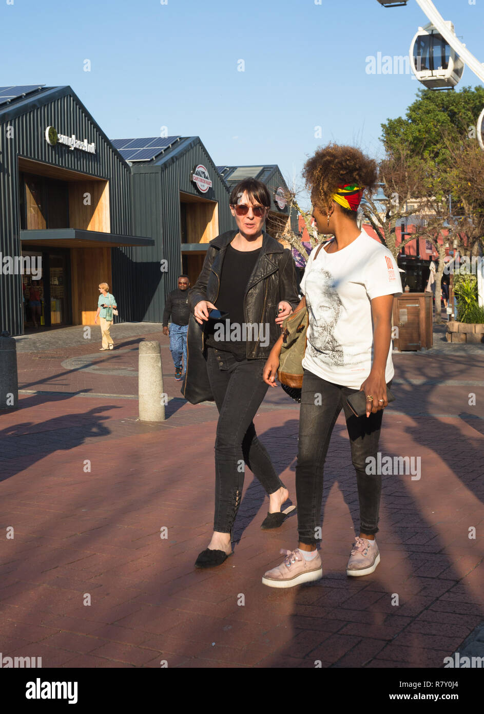 two women female friends and millennials chatting and walking together dressed in fashionable street gear or clothes in the Waterfront in Cape Town Stock Photo