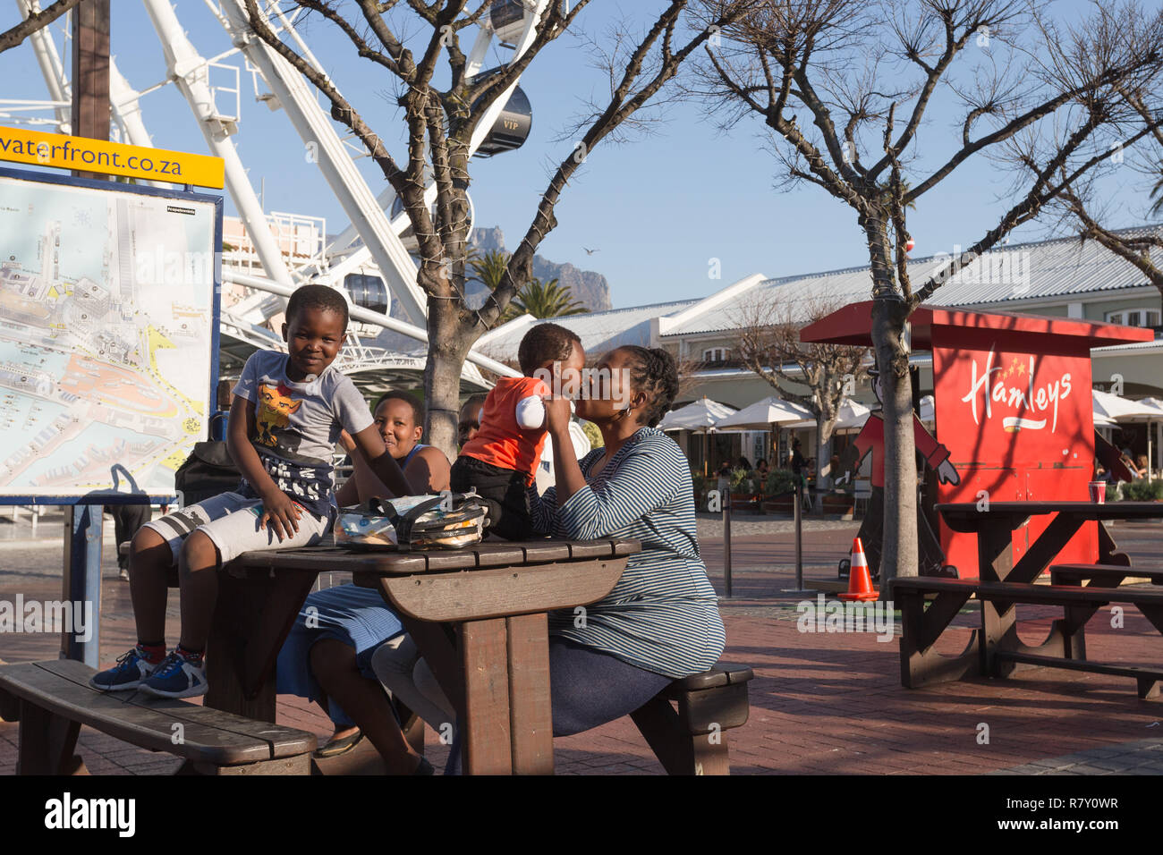 affectionate woman and toddler bonding together seated at a wooden bench outdoors at the V&A Waterfront, Cape Town together with other family members Stock Photo