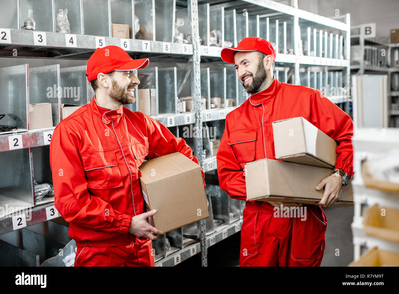 Two handsome warehouse workers in red uniform standing together with boxes in the storage with auto parts or other products Stock Photo