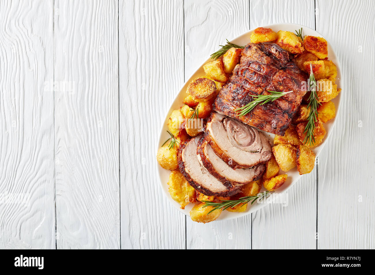 sliced roast pork roulade -  Porchetta, delicious pork roast with crusty roast potatoes served on an oval dish on white wooden table, close-up, flatla Stock Photo