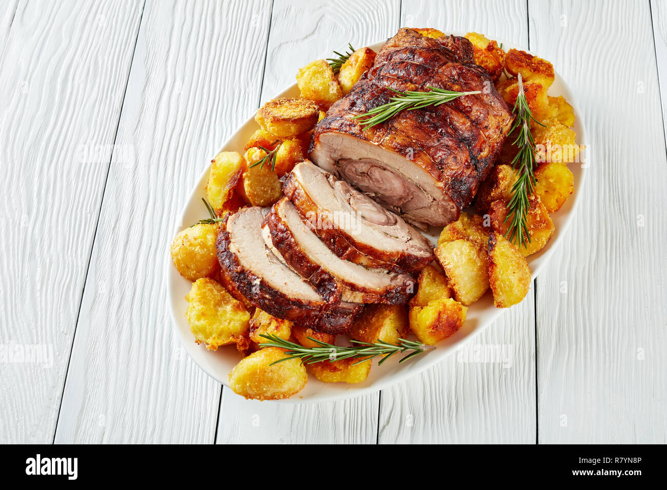 sliced roast pork roulade -  Porchetta, delicious pork roast with crusty roast potatoes served on an oval dish on white wooden table, close-up Stock Photo