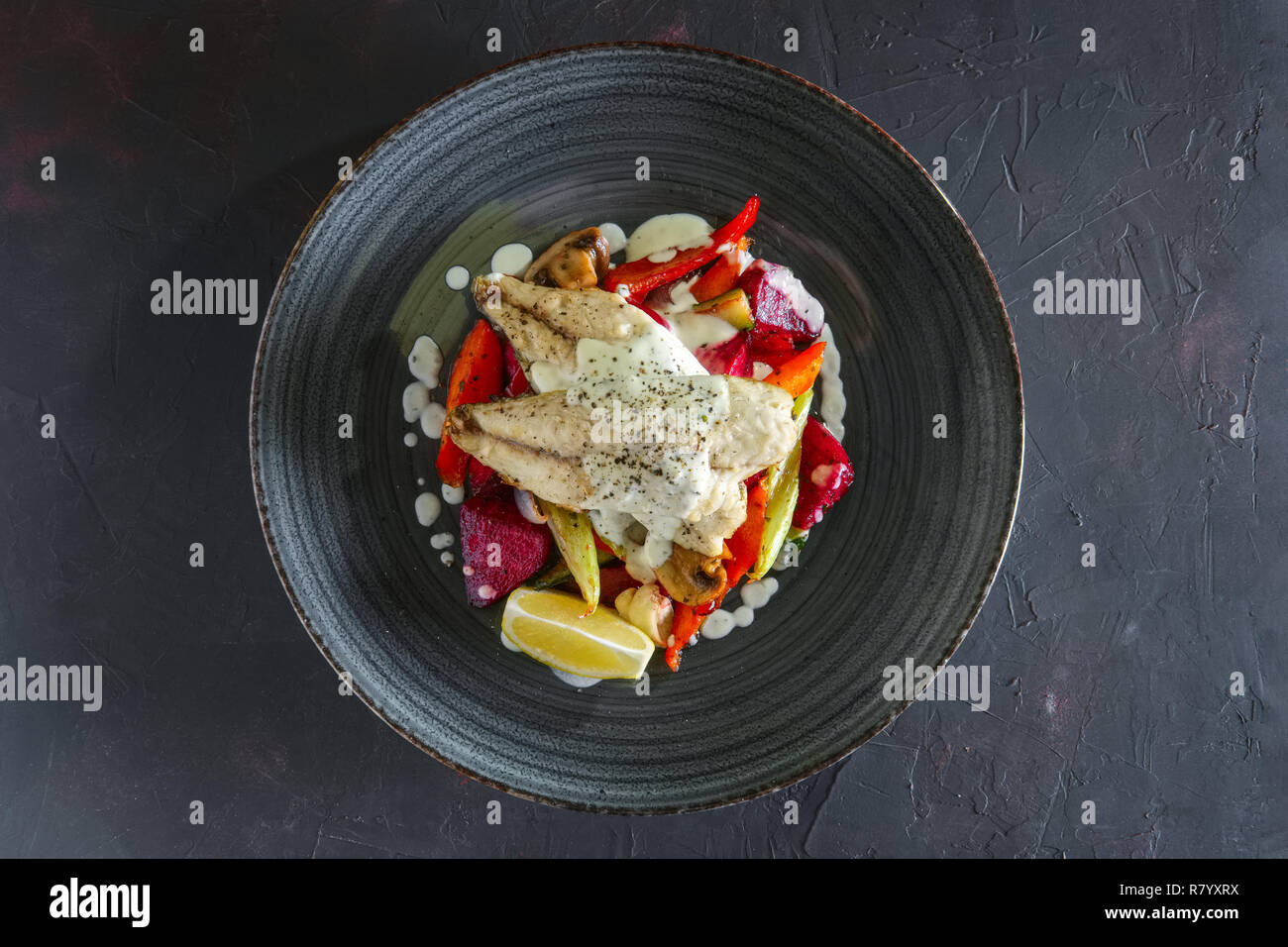Fried fish fillet with grilled beetroot, carrot, bell pepper, zucchini, garlic on dark background Stock Photo