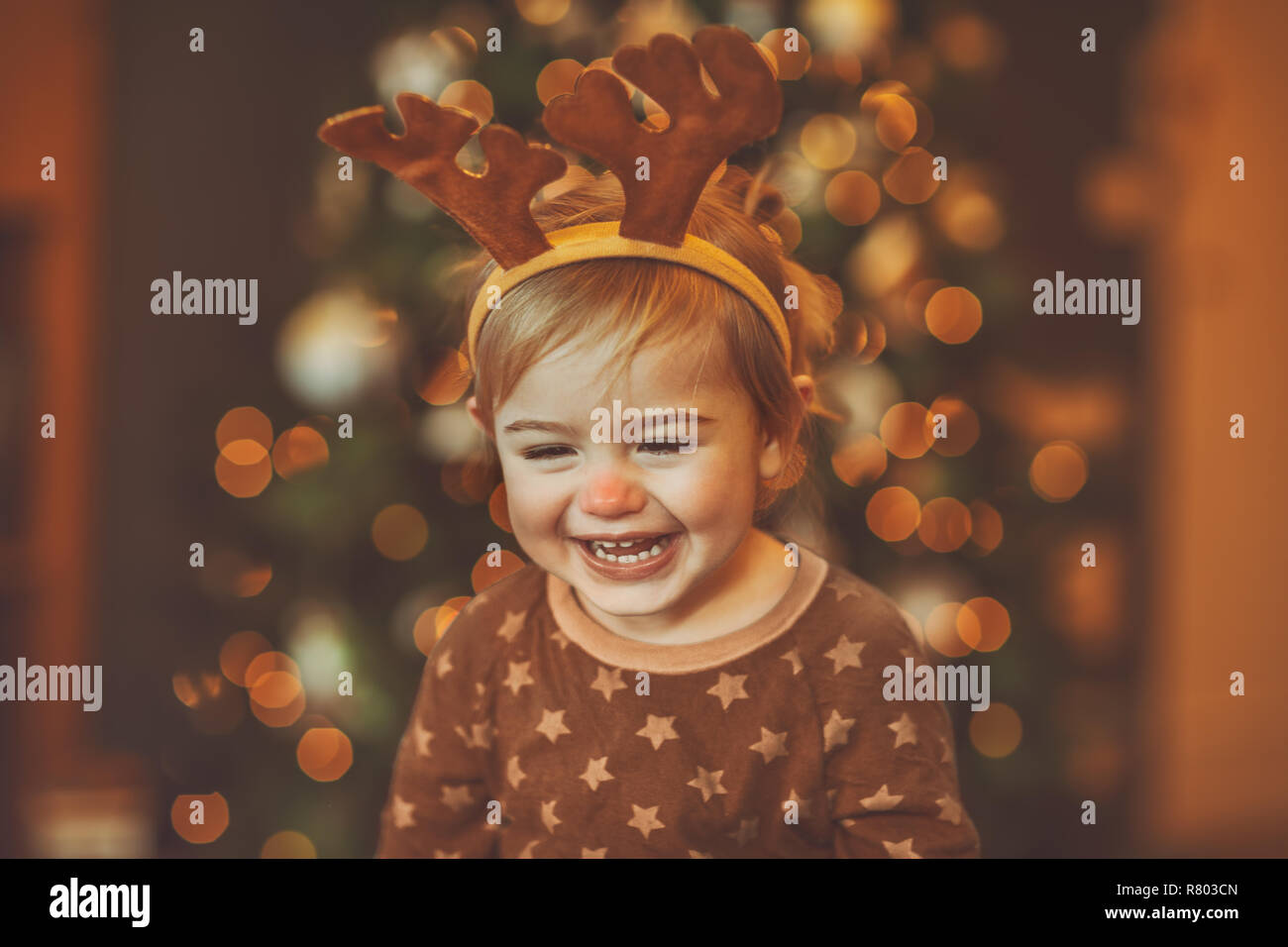 Portrait of a cheerful laughing baby wearing Rudolphs reindeer outfit, with pleasure spending Christmas at home near beautiful glowing Christmas tree Stock Photo