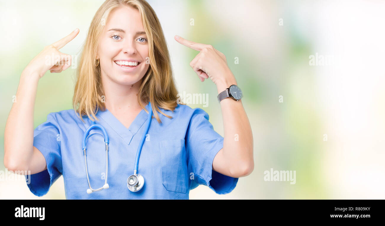 Beautiful young doctor woman wearing medical uniform over isolated background Smiling pointing to head with both hands finger, great idea or thought,  Stock Photo