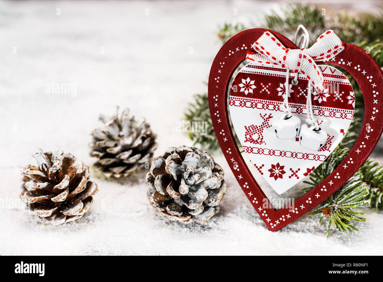 Ornamented wooden heart with Christmas motifs leaning against a fir branch next to a pine cones on the snow. Stock Photo