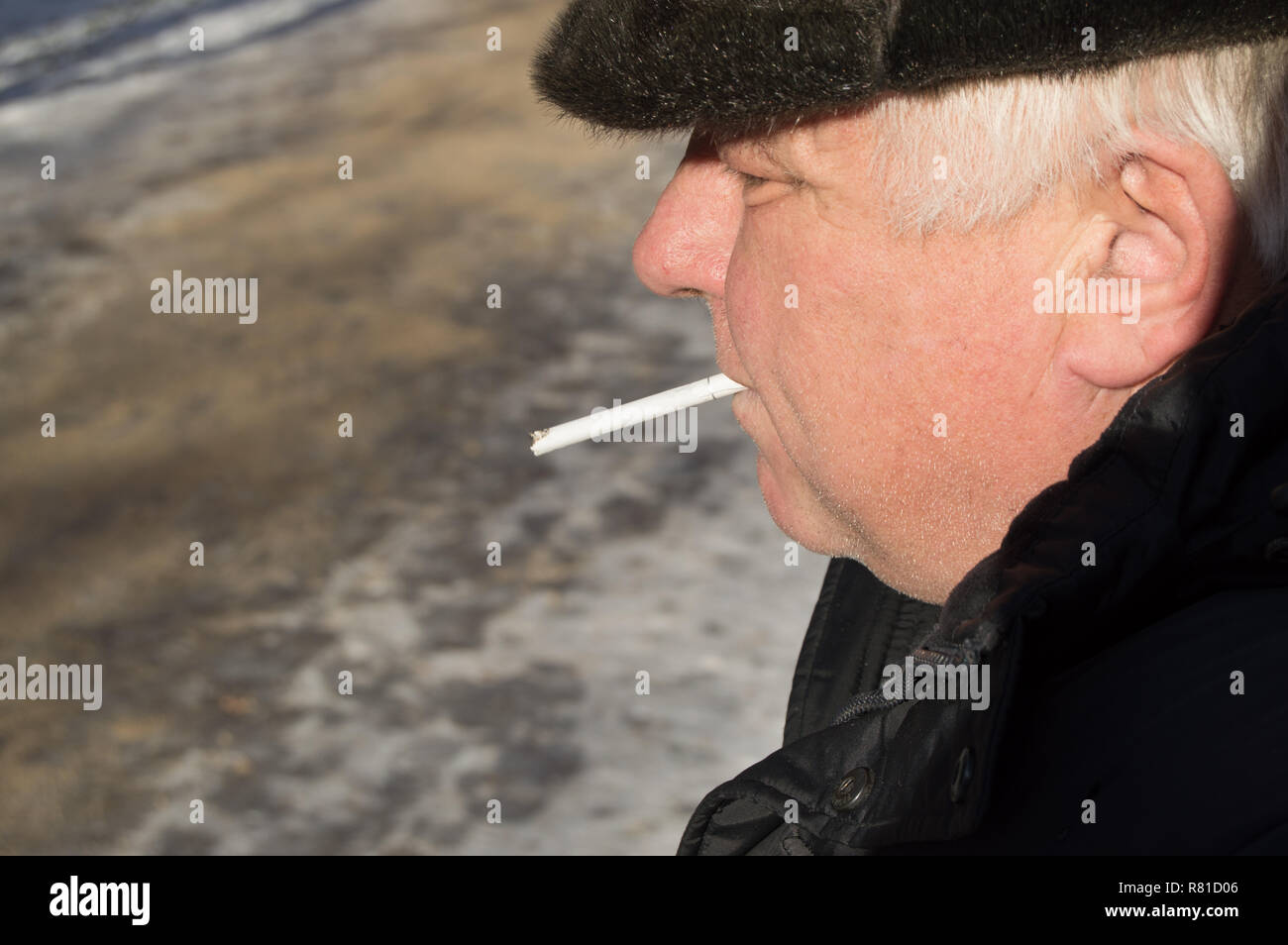 An elderly gray-haired man in winter clothes holding a cigarette in his mouth Stock Photo