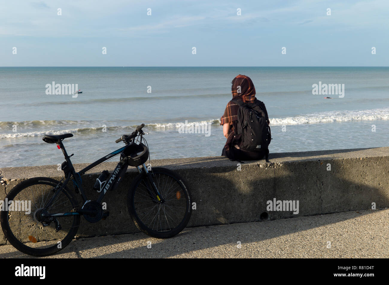 Girl with bicycle sitting on sea wall looking ot to sea, Normandy,France Stock Photo