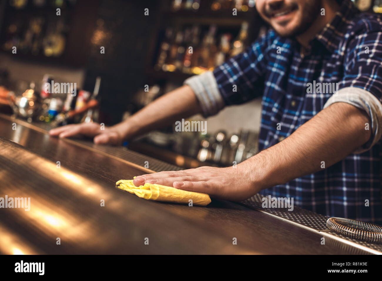 Young bartender standing at bar counter wiping table with rag happy