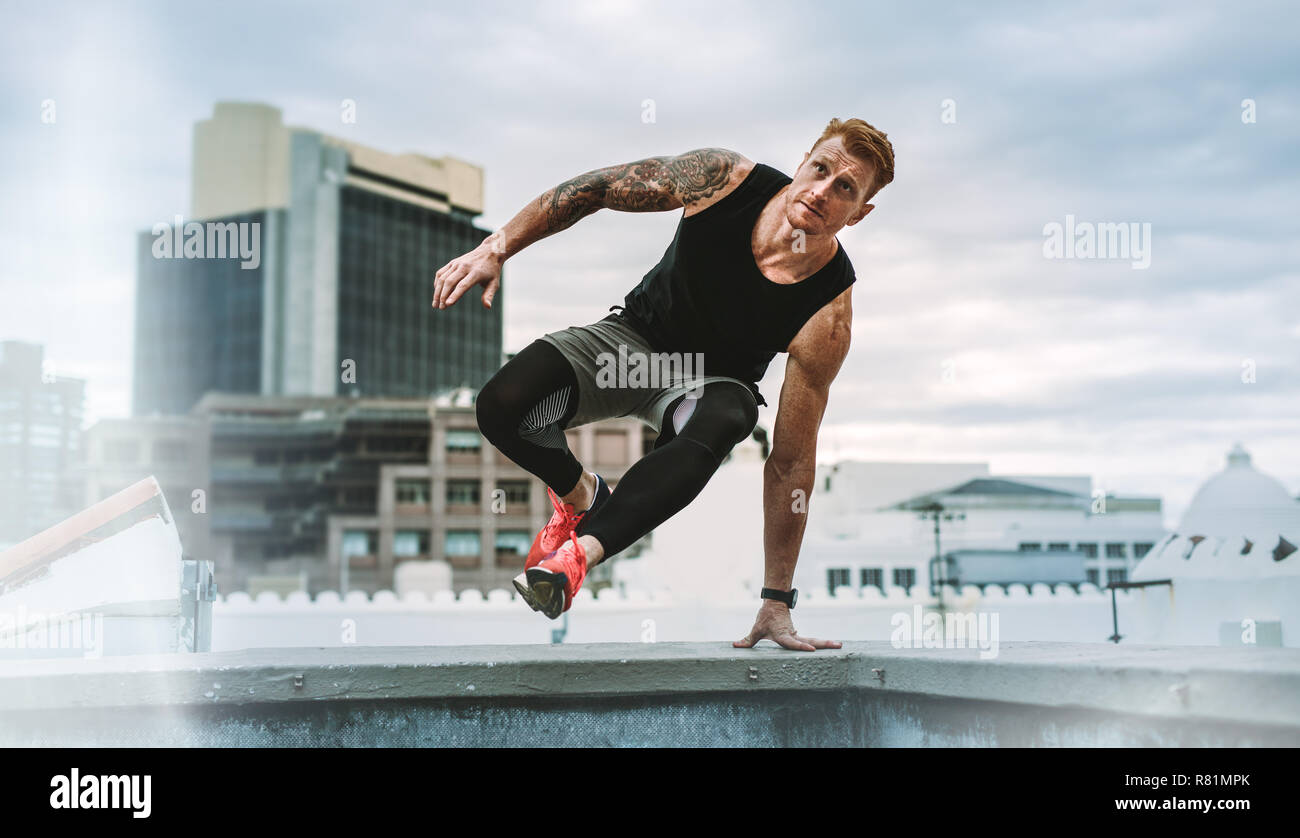 Man doing fitness training on rooftop. Muscular man jumping on to the terrace from the rooftop fence with one hand on fence. Stock Photo