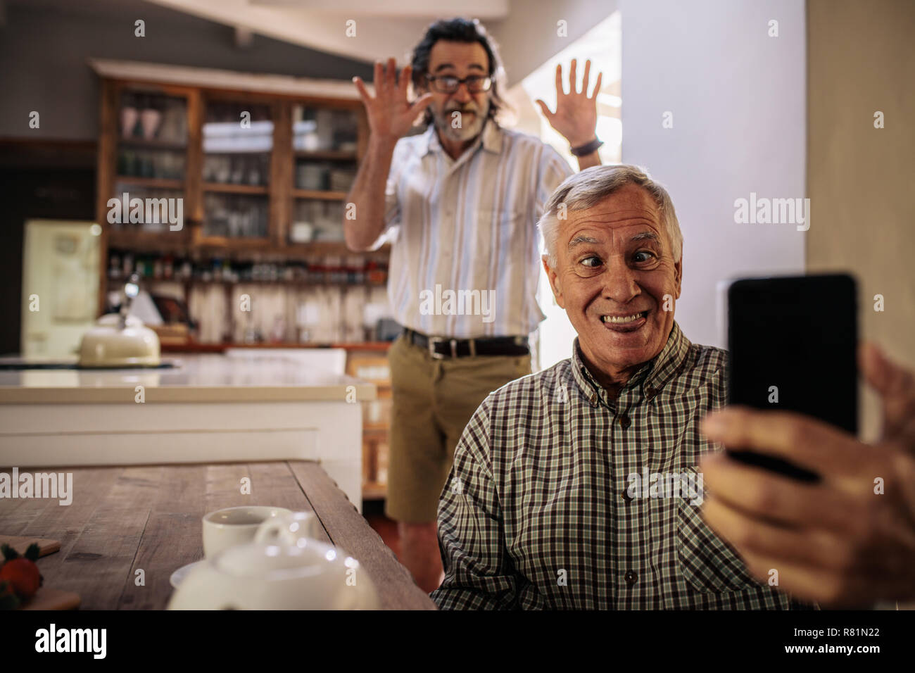 Funny senior man taking selfie with friend standing at back making hand gestures. Retired man making funny face while taking a self portrait with smar Stock Photo