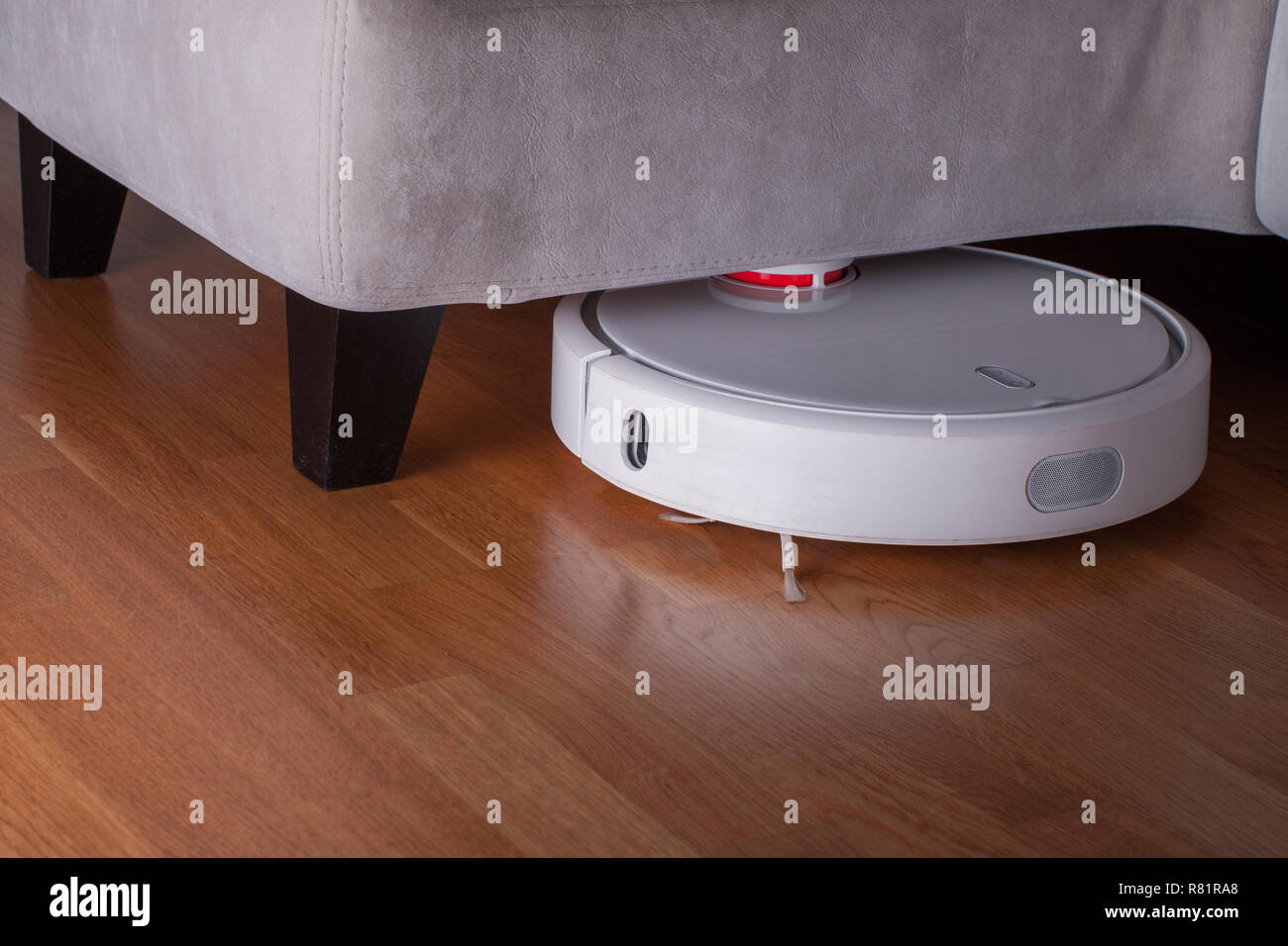 white Robot vacuum cleaner runs in corner under sofa on wood parquet
