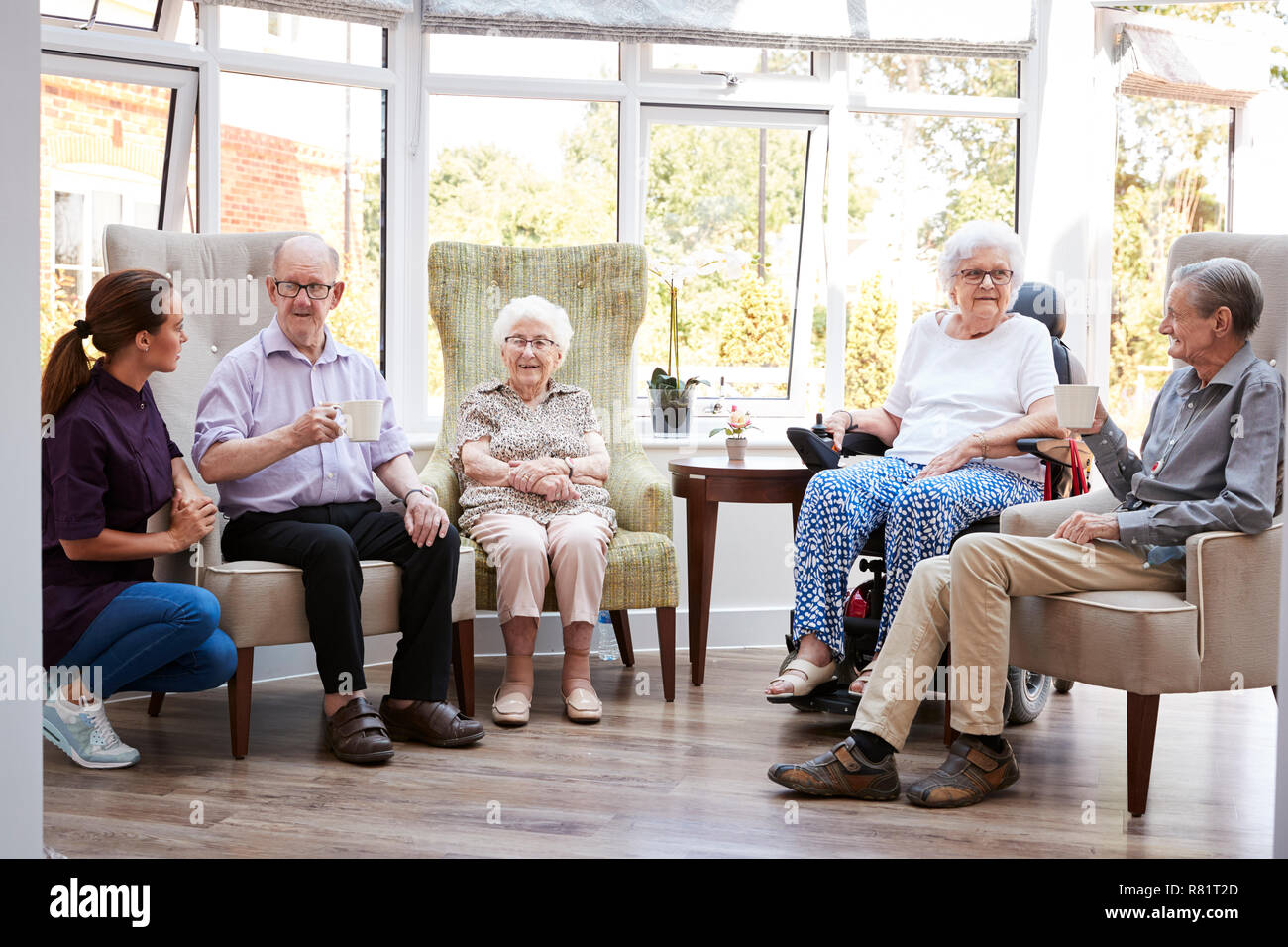 Male And Female Residents Sitting In Chairs And Talking With Carer In Lounge Of Retirement Home Stock Photo