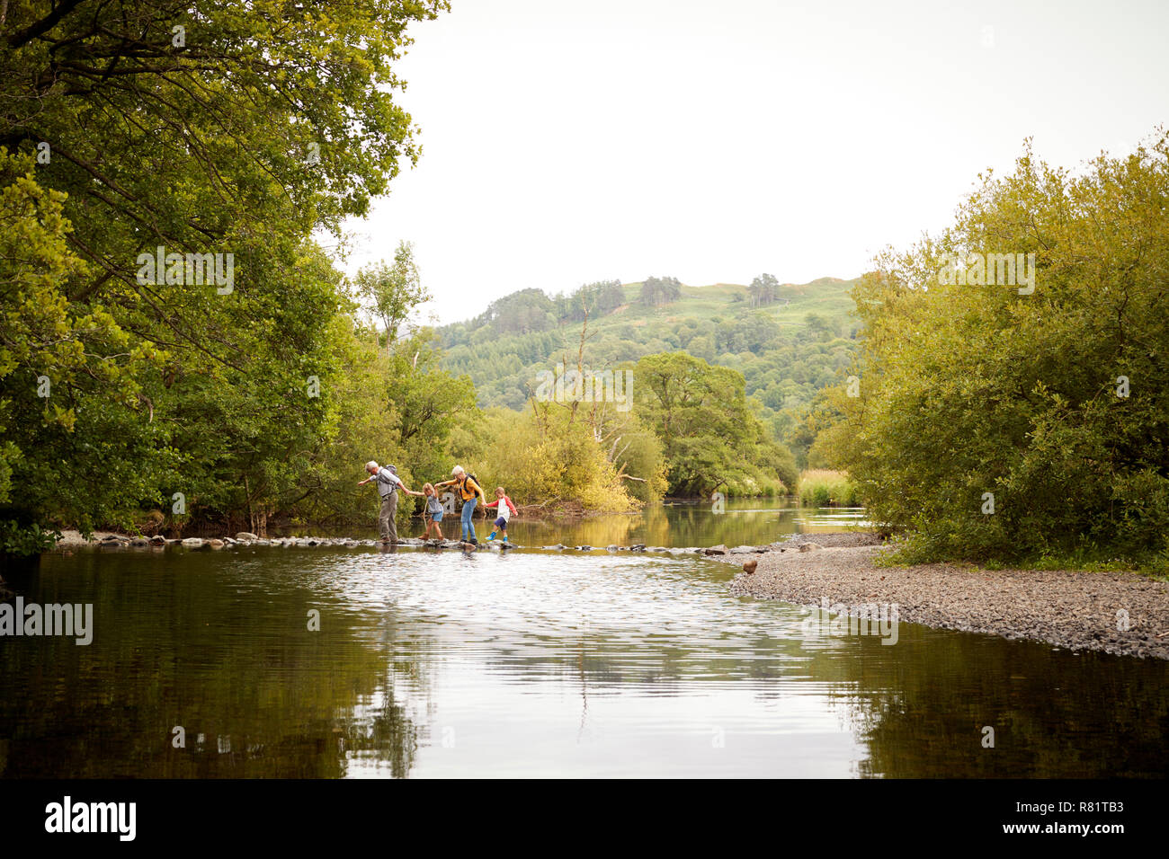 Grandparents With Grandchildren Crossing River Whilst Hiking In UK Lake District Stock Photo