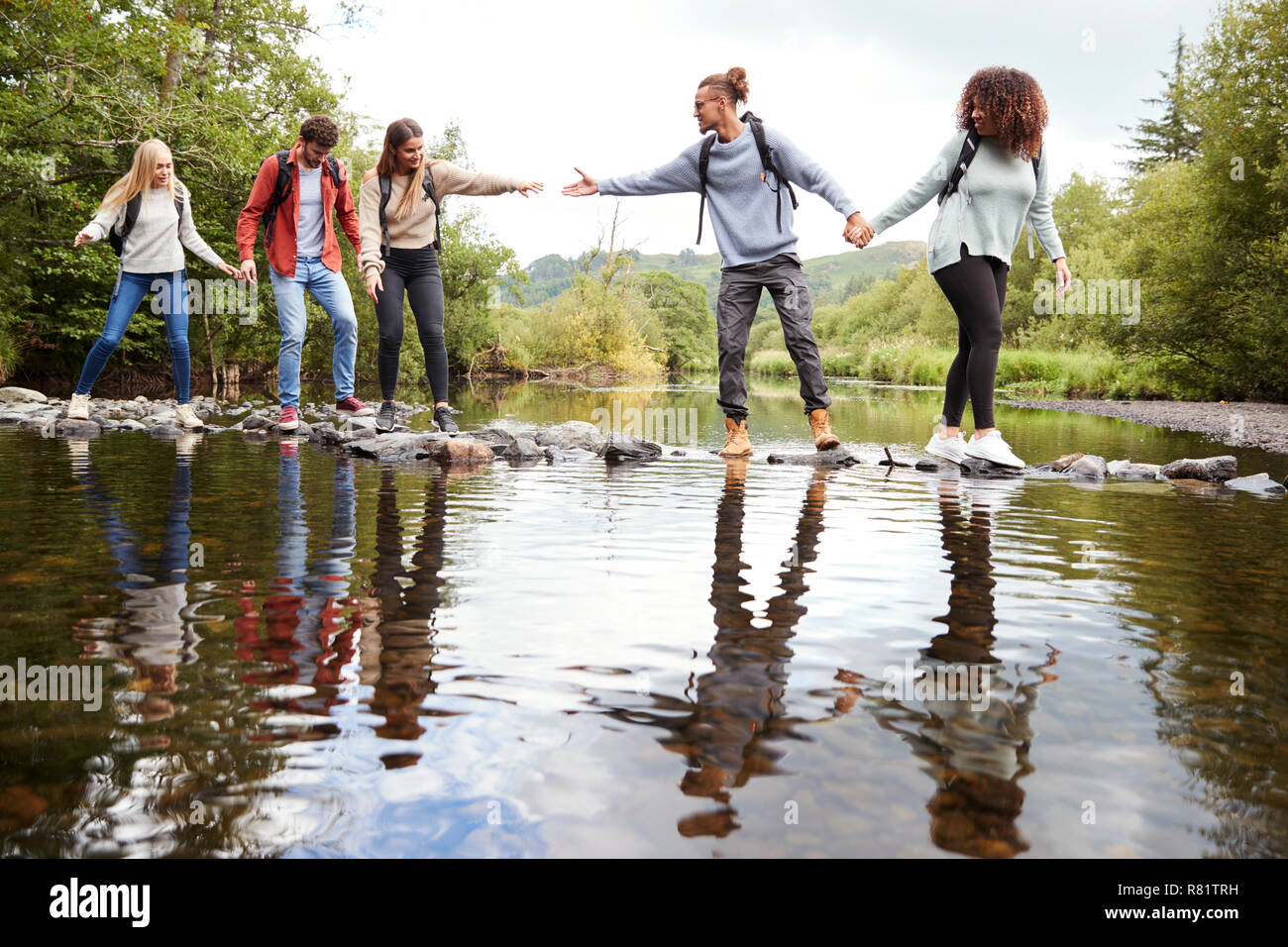 Young adult friends reaching to help each other cross a stream balancing on stones during a hike Stock Photo