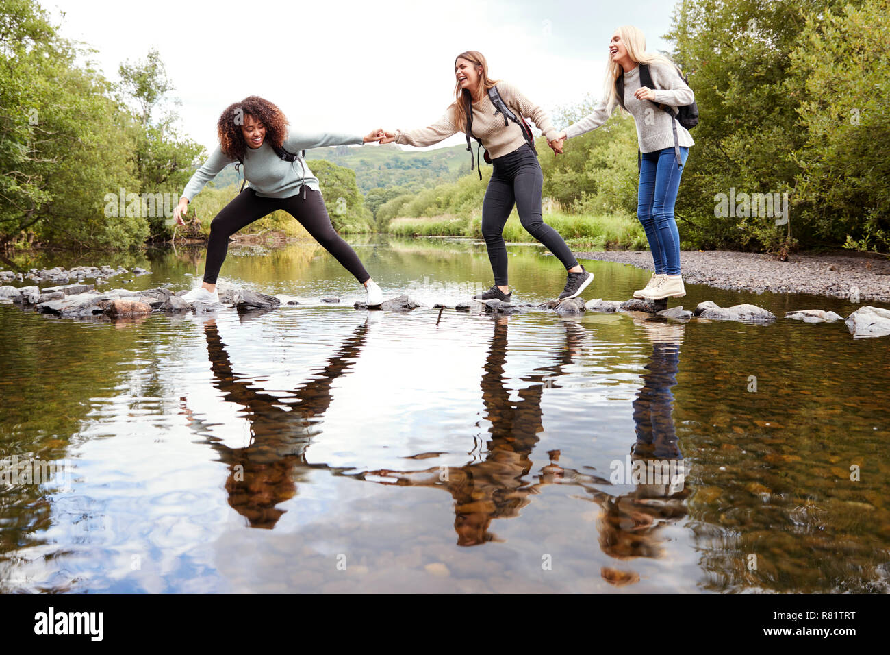 Three young adult women hold hands helping each other while carefully crossing a stream on stones during a hike Stock Photo