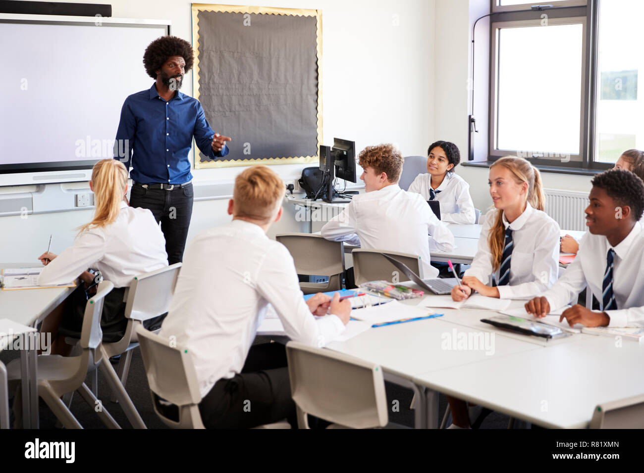 Male High School Teacher Standing Next To Interactive Whiteboard And Teaching Lesson To Pupils Wearing Uniform Stock Photo