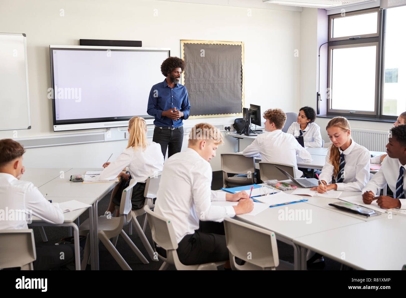 Male High School Teacher Standing Next To Interactive Whiteboard And Teaching Lesson To Pupils Wearing Uniform Stock Photo